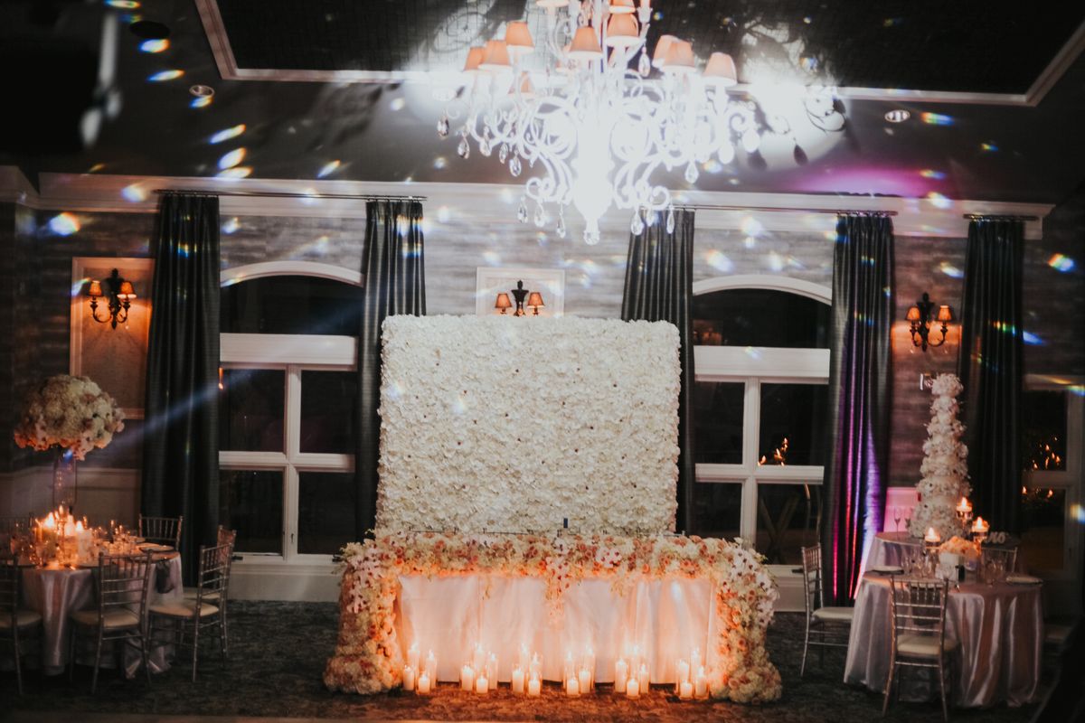Sweetheart table before a white flower wall, glowing with candles